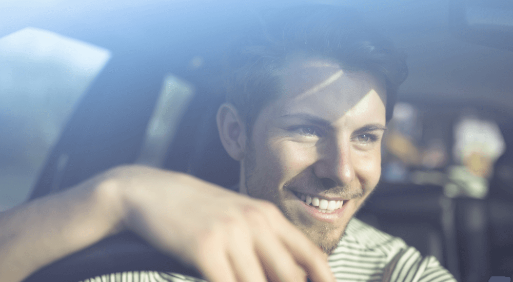 Smiling young man sitting in his car in dappled sunlight with his arm resting over the top of the sterring wheel